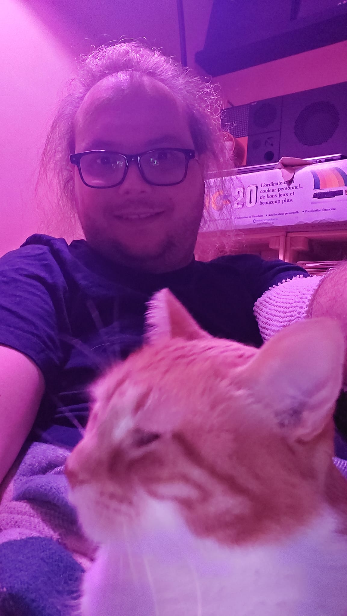 A man with long hair lies on a couch with an orange cat on his lap, the light is purple and other colours are hard to descern